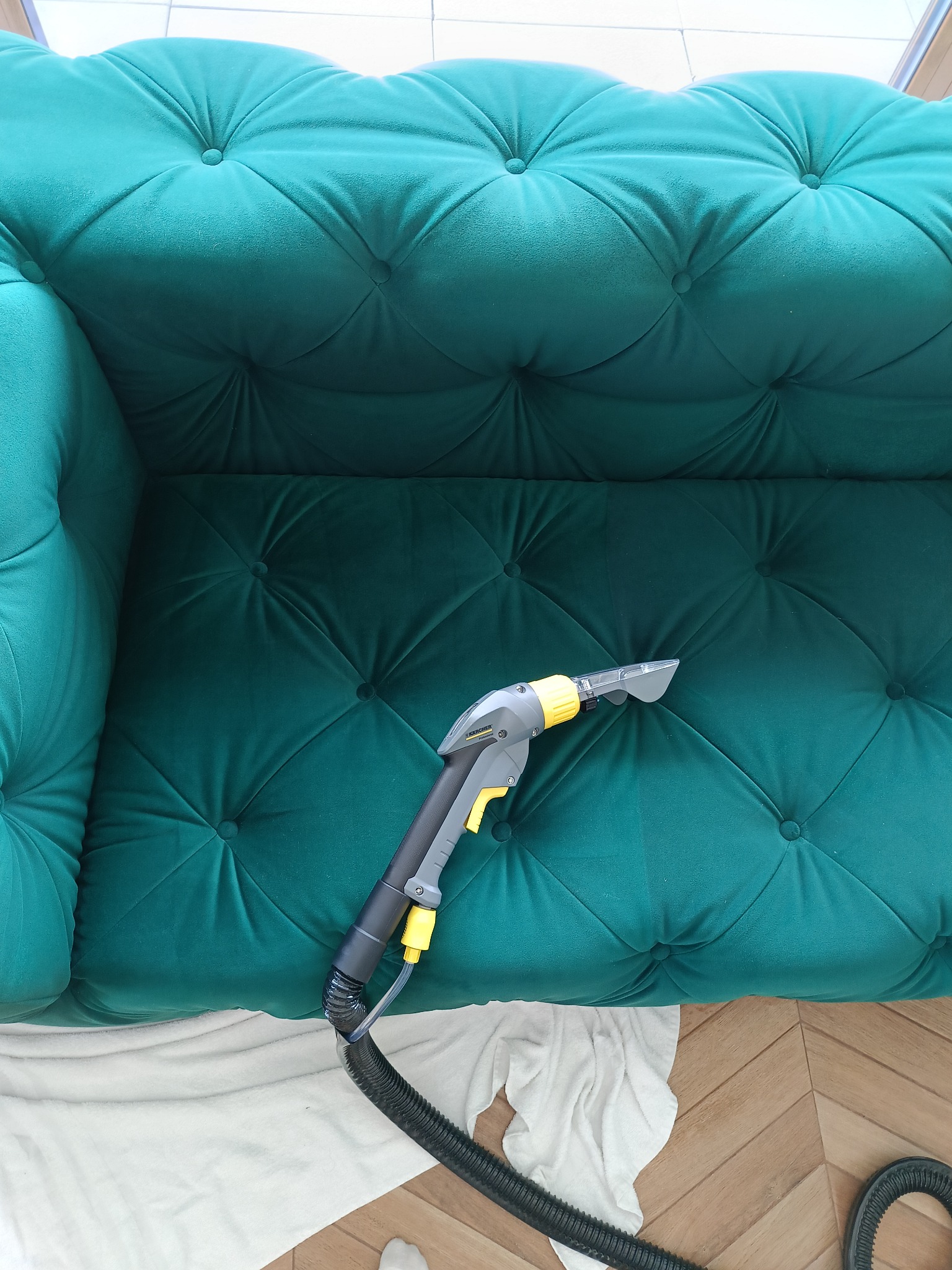 Szczegółowe czyszczenie turkusowej sofy z guzikami za pomocą profesjonalnego odkurzacza piorącego Karcher, na podłodze leży biały ręcznik.