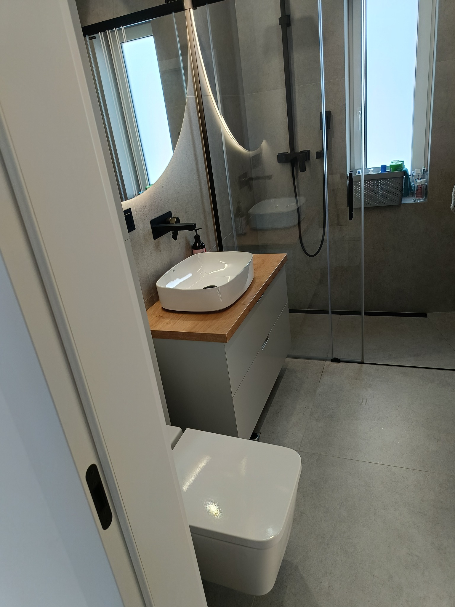 Nowoczesna łazienka w szarościach: kabina prysznicowa ze szkła, umywalka nablatowa z drewnianym blatem, podświetlane lustro i toaleta. Minimalistyczny design.