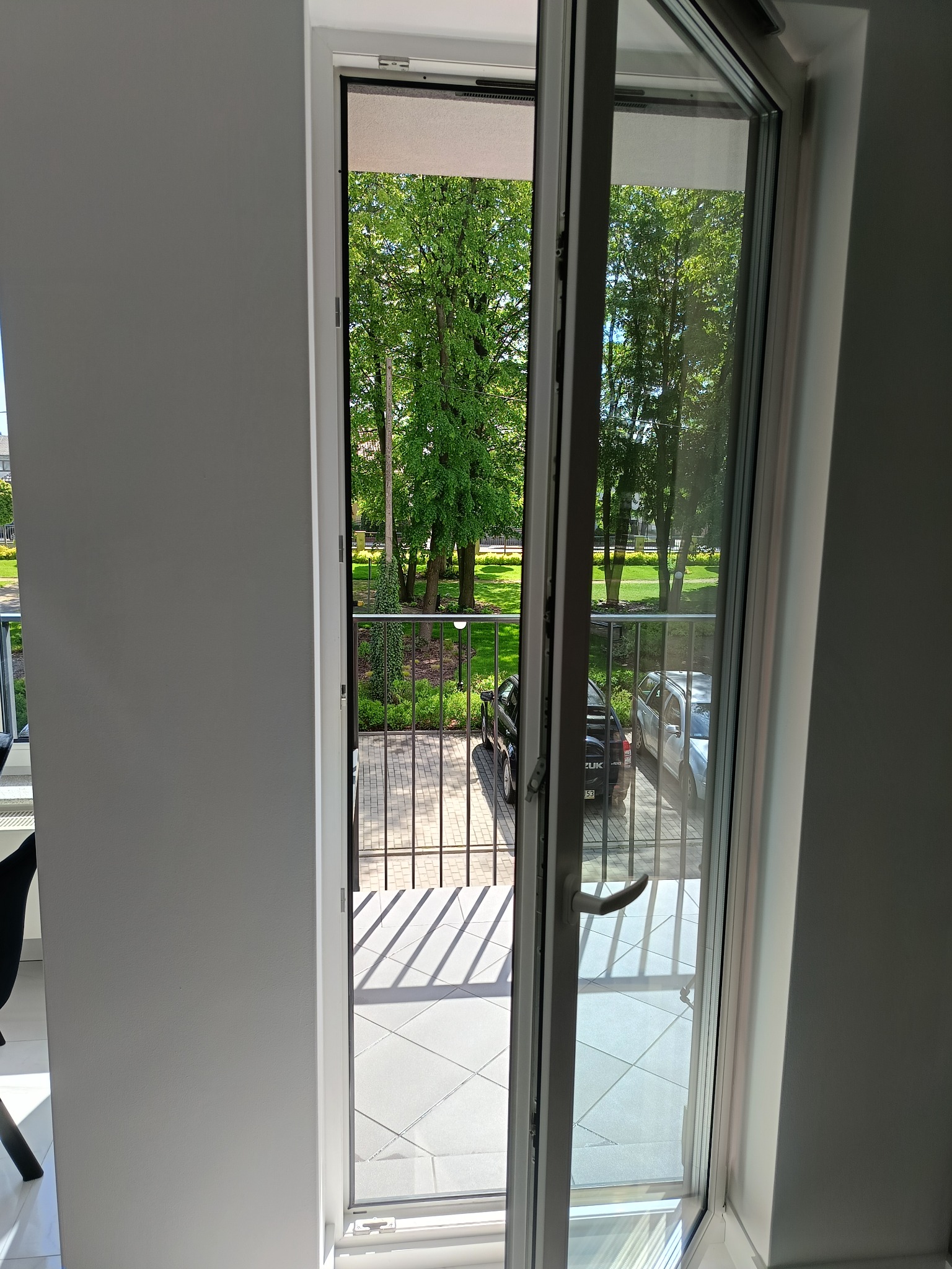 Otwarty balkon z widokiem na park. Białe płytki na podłodze balkonu i metalowa barierka. Za barierką widać zieleń parku i zaparkowane samochody.