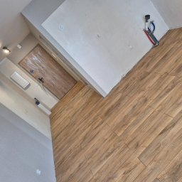 Ł.O. ŁUKASZ ODRZYWOŁEK - Ukończone układanie paneli podłogowych w jasnym wnętrzu. Widoczne drewnopodobne panele, fragment ściany z instalacją hydrauliczną i zabezpieczone folią drzwi.