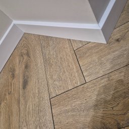 Ł.O. ŁUKASZ ODRZYWOŁEK - Ukończona podłoga z paneli drewnopodobnych, ułożonych w jodełkę, z białą listwą przypodłogową przy ścianie. Widok z góry, bliski kadr.