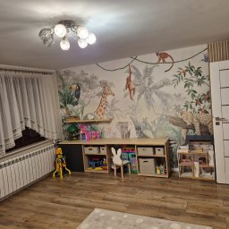 Ł.O. ŁUKASZ ODRZYWOŁEK - Pokój dziecięcy z fototapetą w motywy dżungli, drewnianą podłogą i meblami do przechowywania zabawek. Widoczne okno z firanką i biały grzejnik.