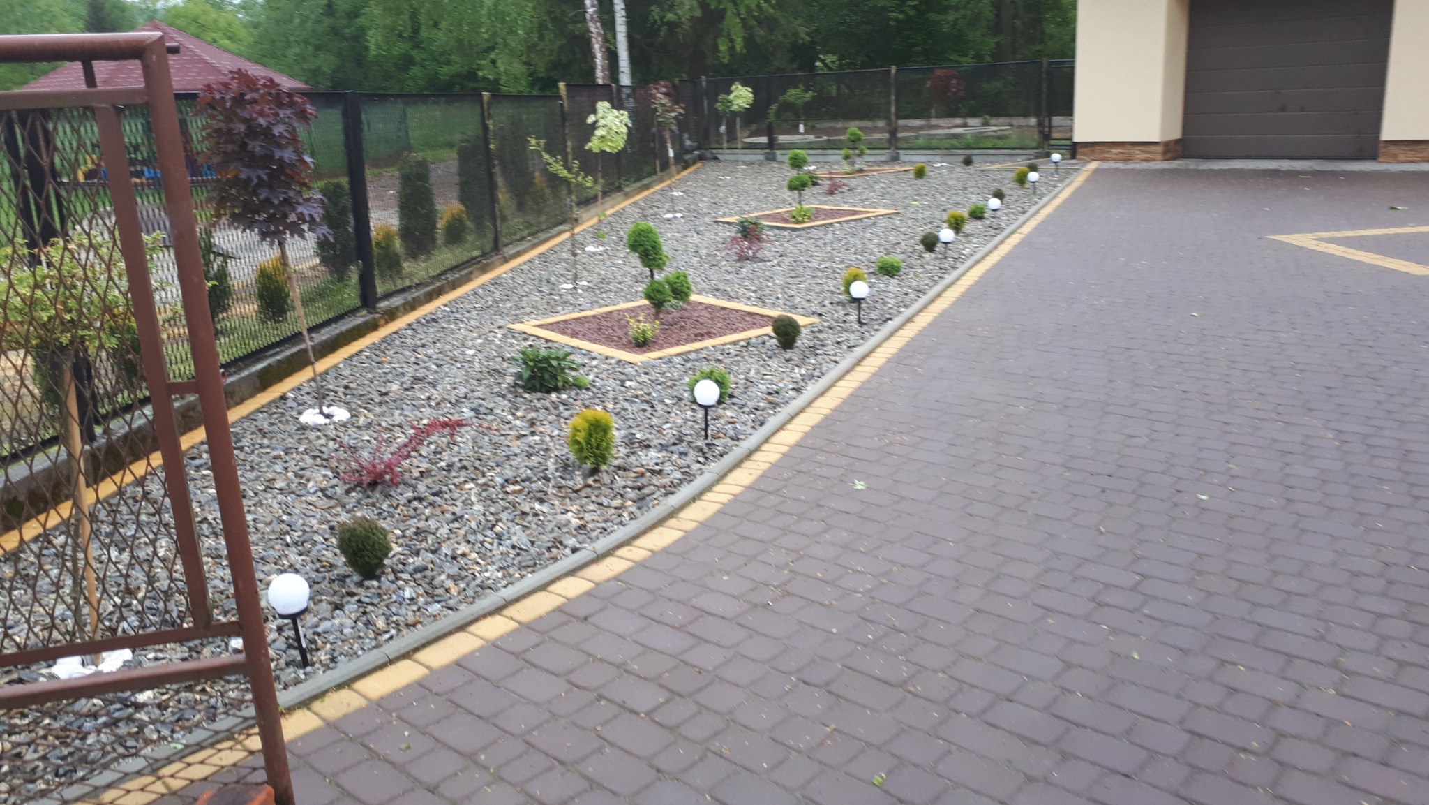 Uporządkowany ogród dekoracyjny z nasadzeniami w żwirze, oświetlony kulistymi lampami, widok z brukowanego podjazdu.