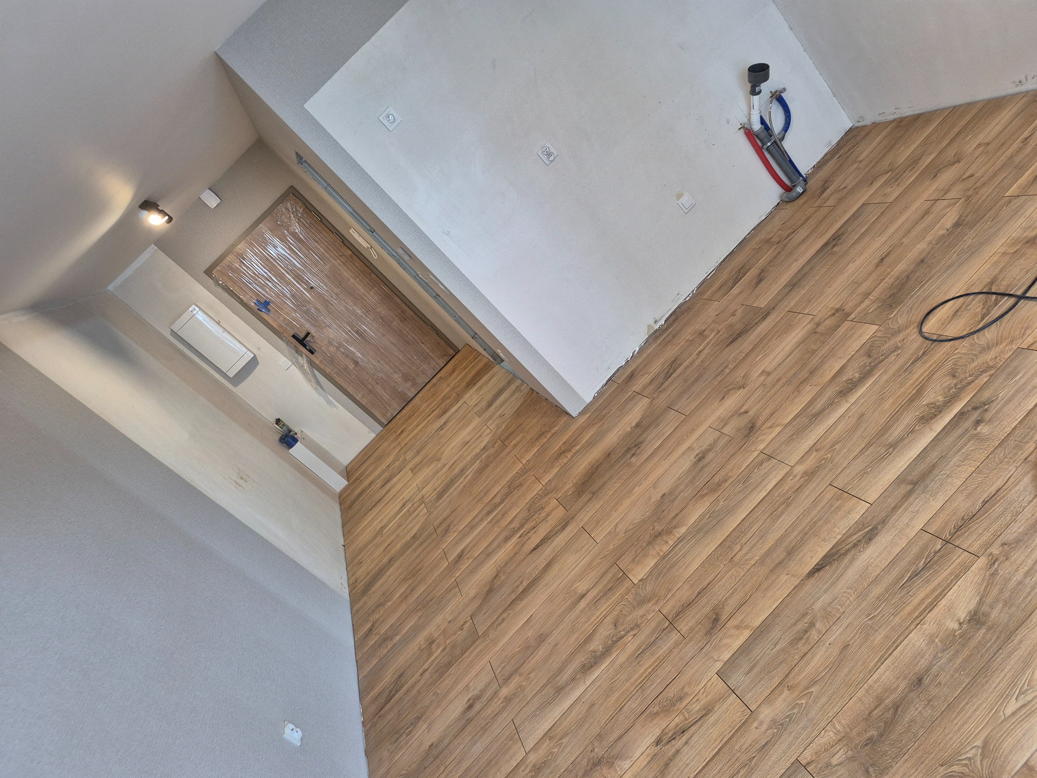 Ukończone układanie paneli podłogowych w jasnym wnętrzu. Widoczne drewnopodobne panele, fragment ściany z instalacją hydrauliczną i zabezpieczone folią drzwi.