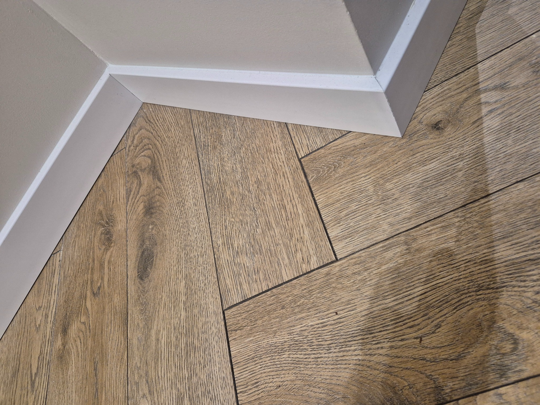 Ukończona podłoga z paneli drewnopodobnych, ułożonych w jodełkę, z białą listwą przypodłogową przy ścianie. Widok z góry, bliski kadr.