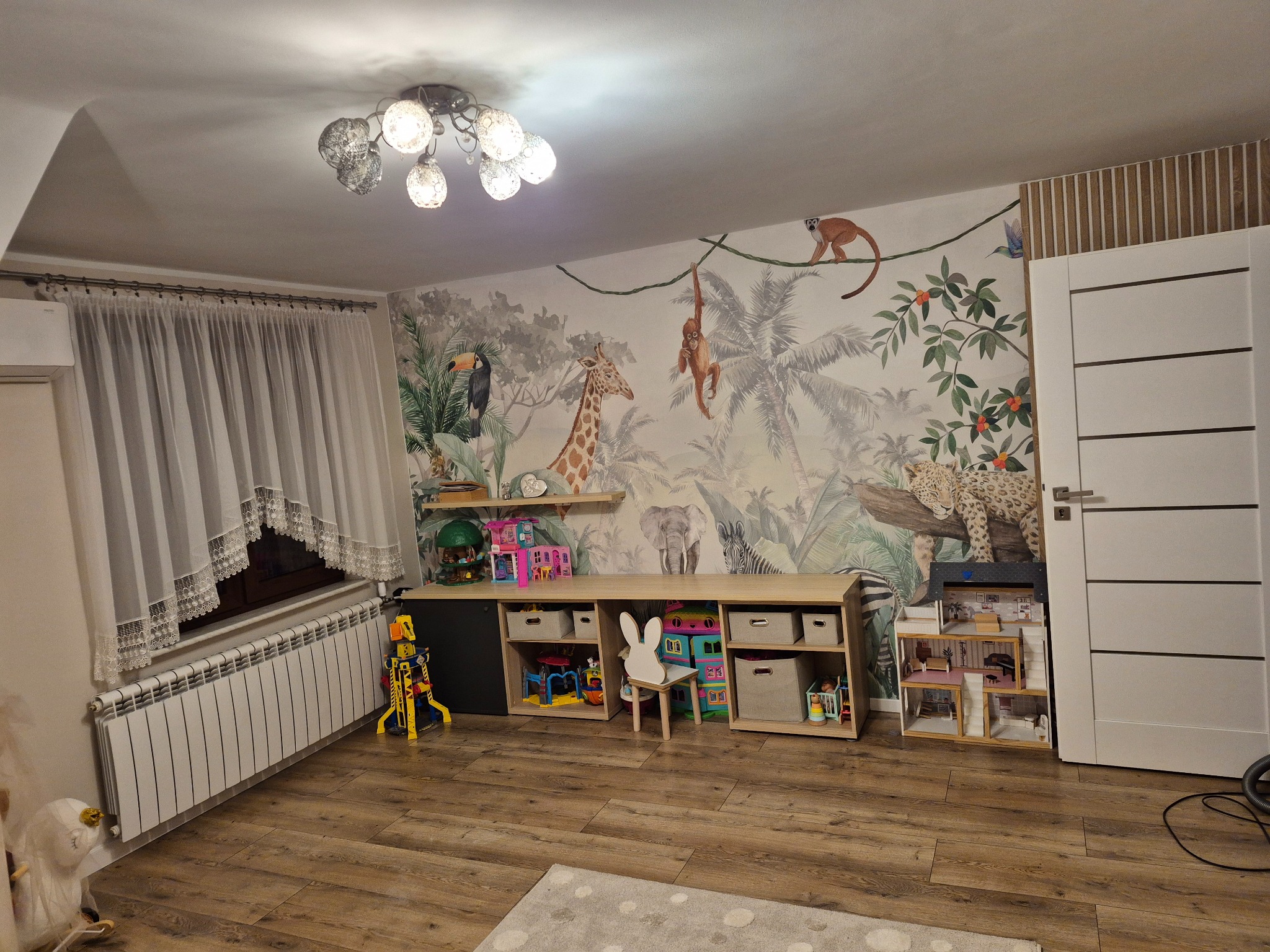 Pokój dziecięcy z fototapetą w motywy dżungli, drewnianą podłogą i meblami do przechowywania zabawek. Widoczne okno z firanką i biały grzejnik.