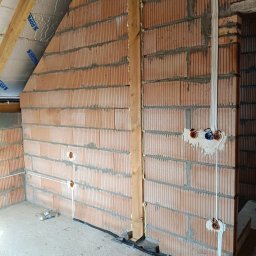 Wiktor Hanowicz - Wnętrze w trakcie prac wykończeniowych: murowana ściana z cegły, widoczne przewody elektryczne zamontowane natynkowo, puszki pod gniazdka i włączniki w ścianie, drewniany słup podtrzymujący strop...