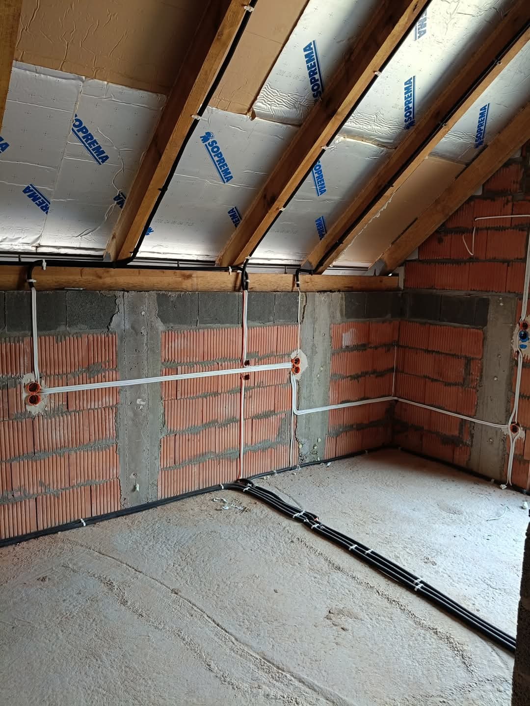 Wnętrze w trakcie prac wykończeniowych: widoczne ceglane ściany z rozprowadzoną instalacją elektryczną, białe kable w peszlach, puszki pod gniazdka i włączniki, oraz czarne rury instalacyjne...