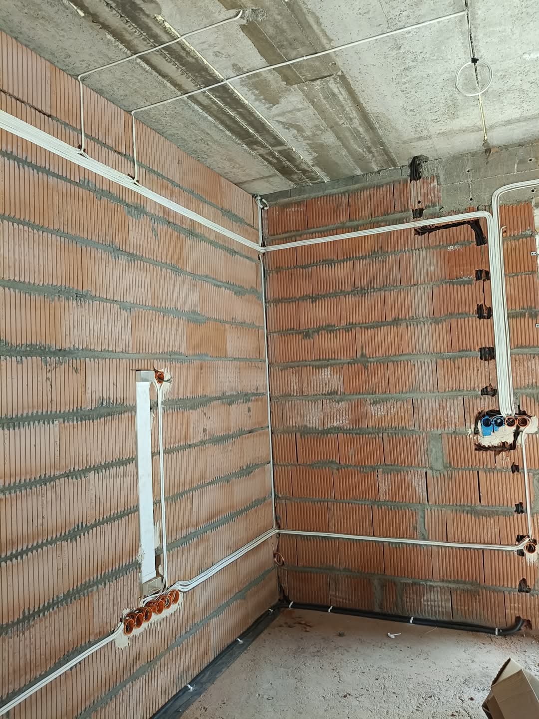 Surowe wnętrze w trakcie prac: widoczne ceglane ściany z rozprowadzonymi białymi przewodami elektrycznymi, gdzieniegdzie puszki pod gniazdka i wyprowadzone przewody, surowy betonowy sufit.