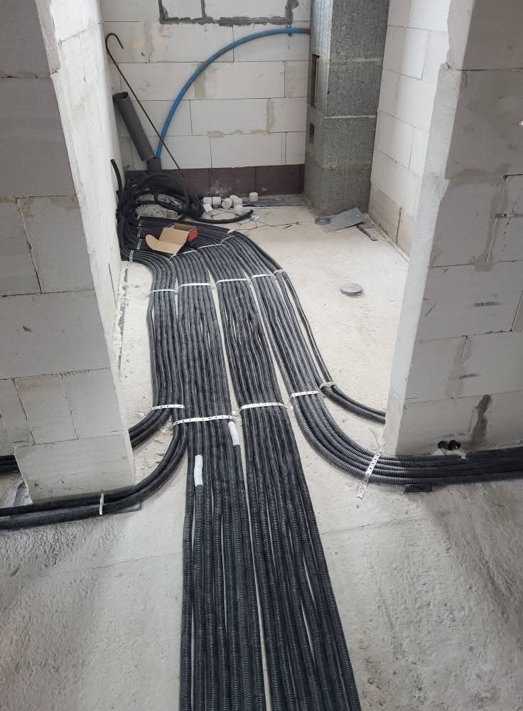 Wiązki przewodów elektrycznych w peszlach ułożone równolegle na surowym podłożu, w trakcie instalacji wewnątrz budynku z surowymi ścianami z betonu komórkowego.