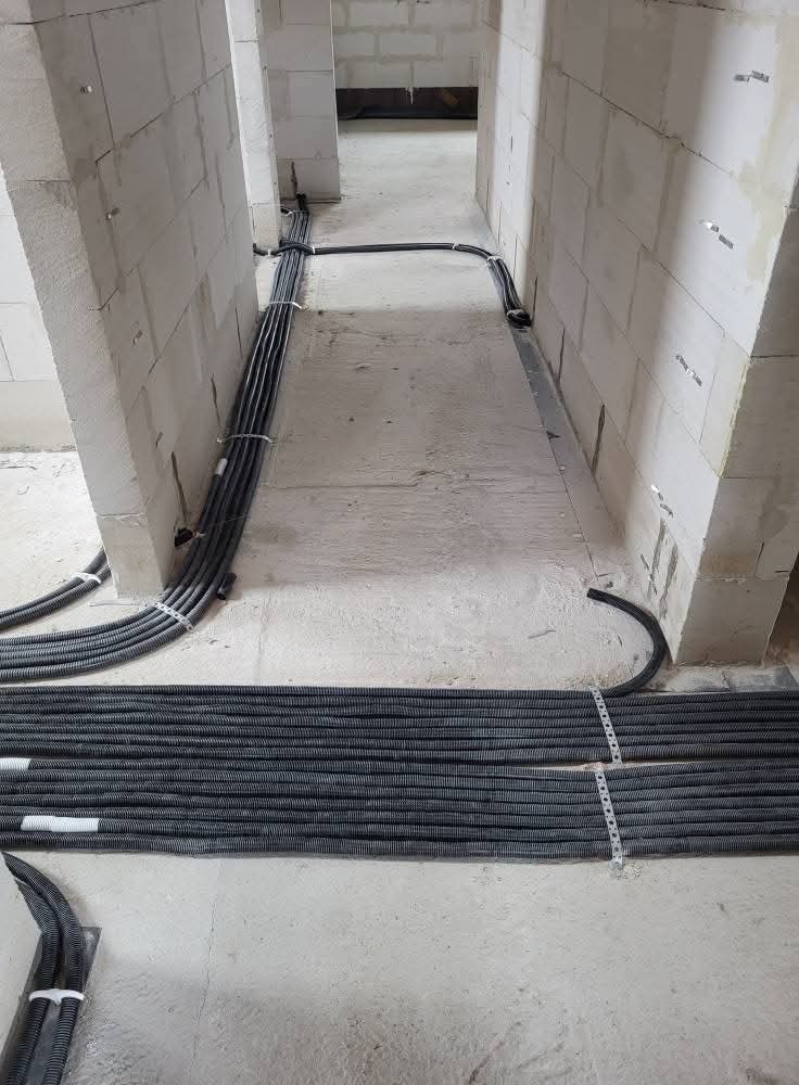 Korytarz w surowym stanie z ułożonymi na podłodze rurami karbowanymi i przewodami elektrycznymi, prowadzącymi wzdłuż ścian z białego bloczka.