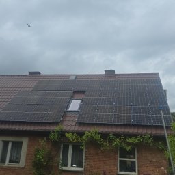 Klaudia Krzyż - Ceglany dom z panelami słonecznymi na brązowym dachu, widok z dołu. Na niebie widoczny ptak. Z boku roślinność.