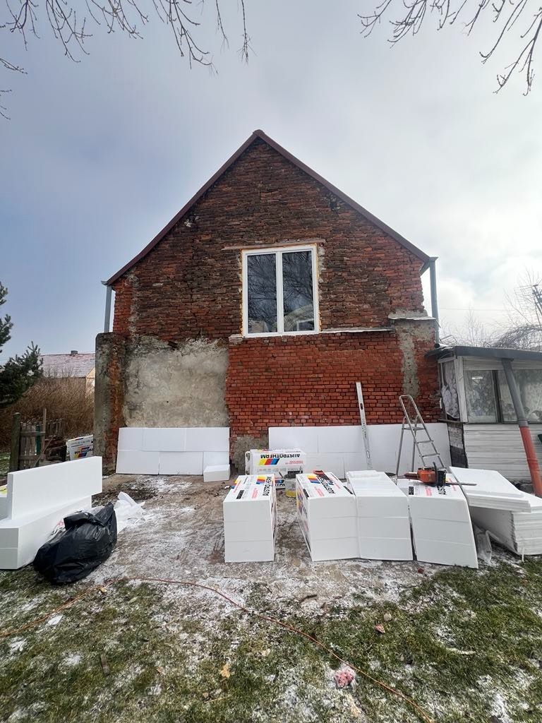 Dom z czerwonej cegły przed termomodernizacją, widoczne przygotowane płyty styropianowe na trawie, drabina oparta o ścianę i częściowo ocieplona ściana z oknem.