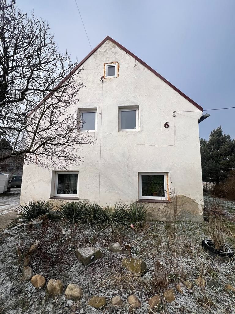 Dwukondygnacyjny dom z numerem 6 na elewacji, widoczny przed termomodernizacją, z fragmentami odpadającego tynku u dołu ściany i oknem zamurowanym na poddaszu, otoczony zaniedbanym ogrodem...