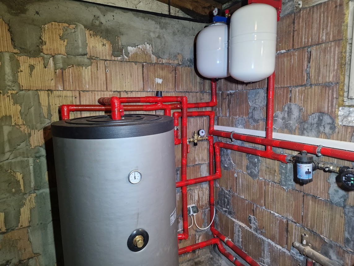 Szary zbiornik z manometrem i czerwone rury w kotłowni z surową cegłą, instalacja grzewcza z dwoma białymi zbiornikami wyrównawczymi.