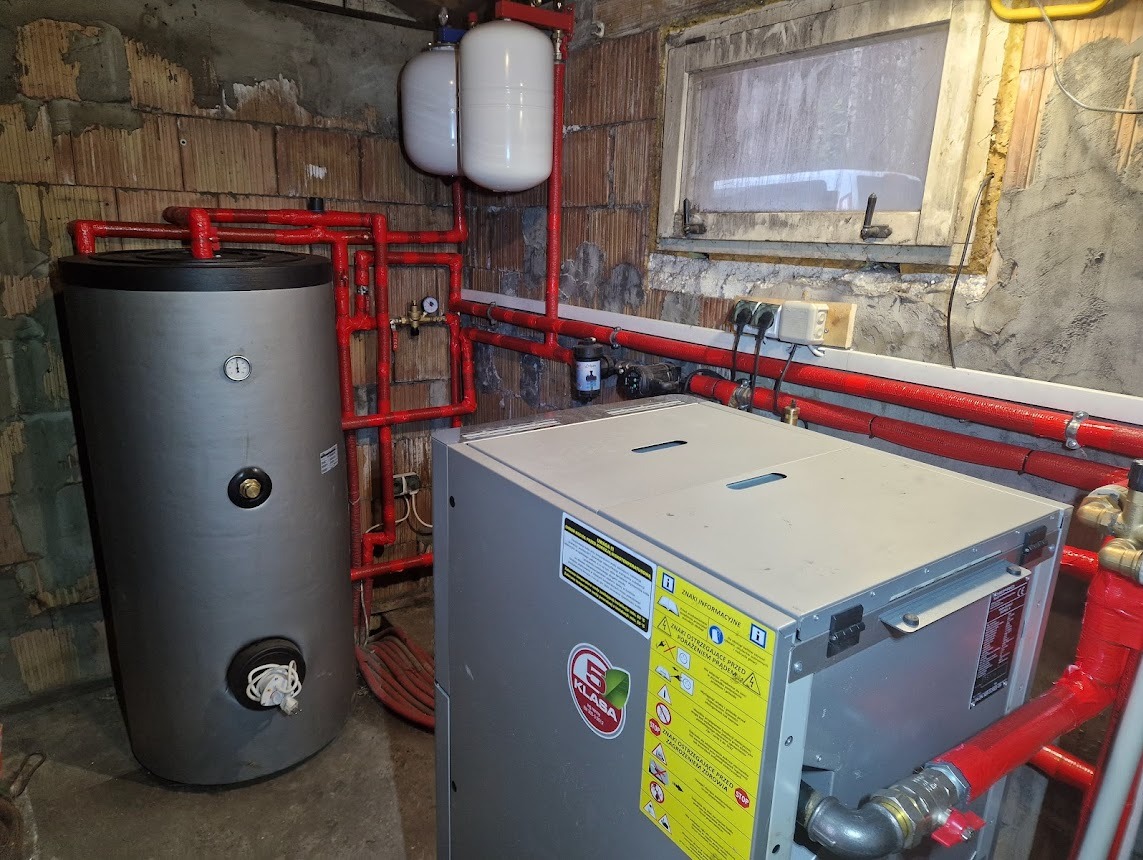 Instalacja z szarym zbiornikiem, czerwonymi rurami i białymi zbiornikami wyrównawczymi w kotłowni z oknem i ceglanymi ścianami. Widoczne elementy instalacji.