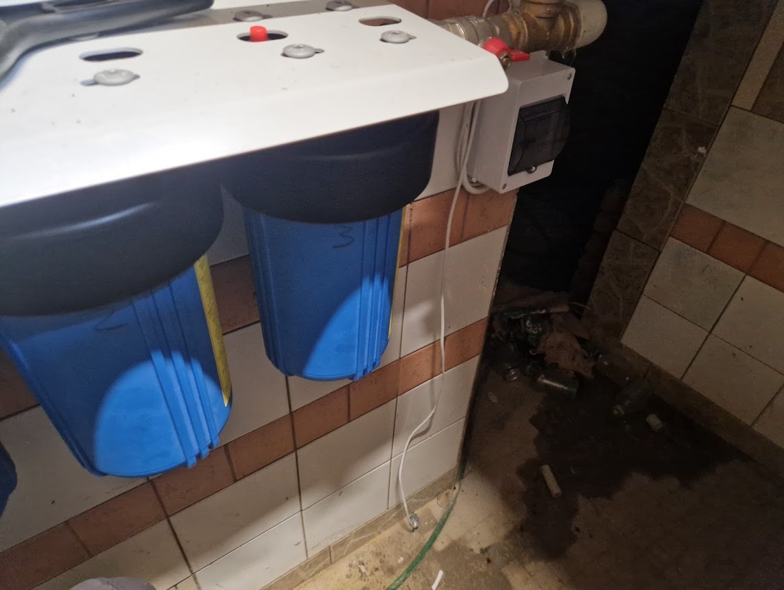 Filtr wody z dwoma niebieskimi zbiornikami, zamontowany na ścianie z brązowymi płytkami. Widoczny przewód i elementy instalacji.