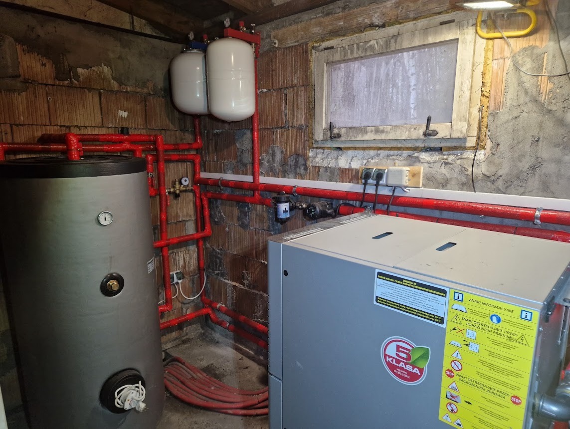 Instalacja zbiornika buforowego z rurami w izolacji w Błotnicy. Widoczny szary zbiornik, białe zasobniki i czerwone rury. Fragment okna w tle.