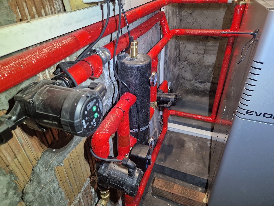 Instalacja grzewcza z czerwonymi rurami i pompami obiegowymi, widoczny zbiornik wyrównawczy i fragment kotła. Elementy zamontowane przy starej ścianie.