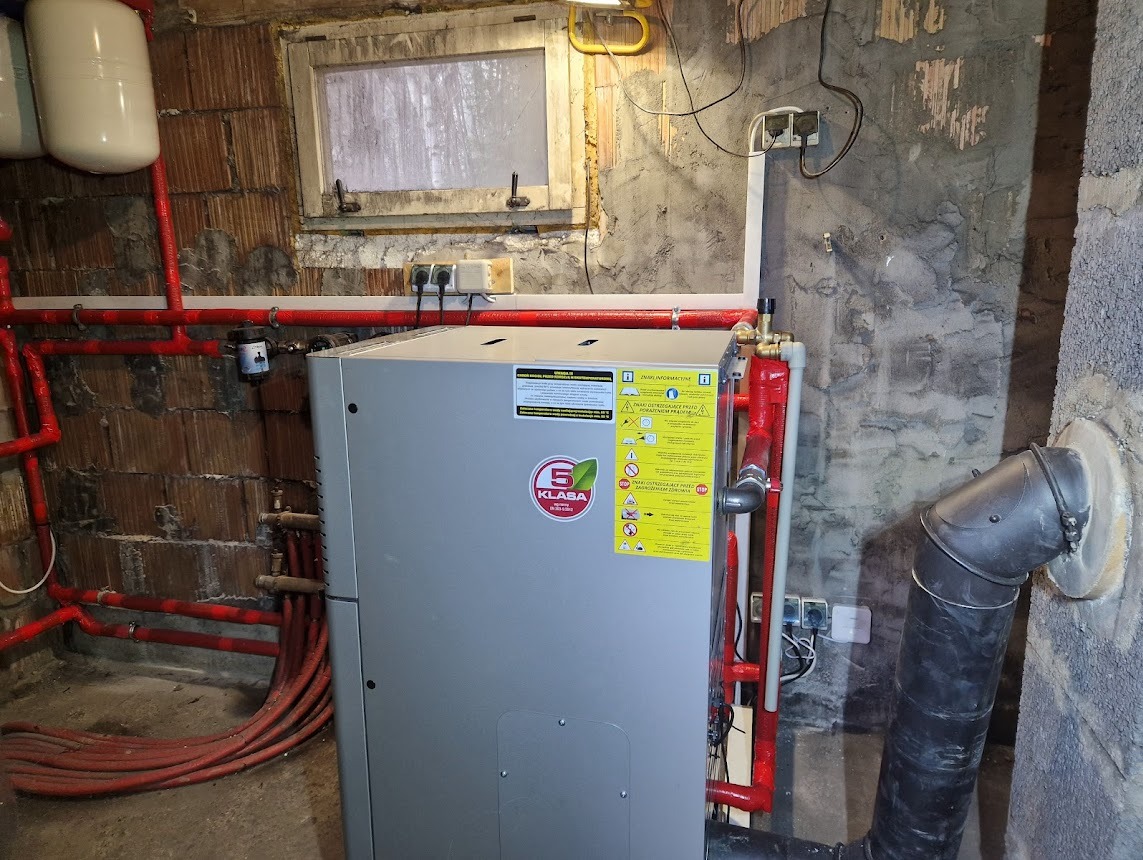 Szara pompa ciepła w piwnicy z instalacją grzewczą. Czerwone rury, okno w tle i surowy beton na ścianach. Widoczne przyłącza i etykiety informacyjne.