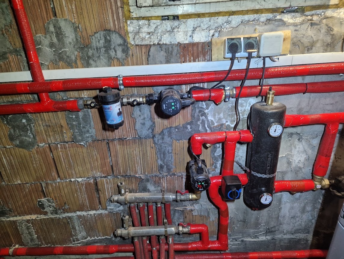 Instalacja grzewcza z czerwonymi rurami, pompą obiegową i filtrem, zamontowana na surowej, ceglanej ścianie z widocznymi przewodami elektrycznymi.