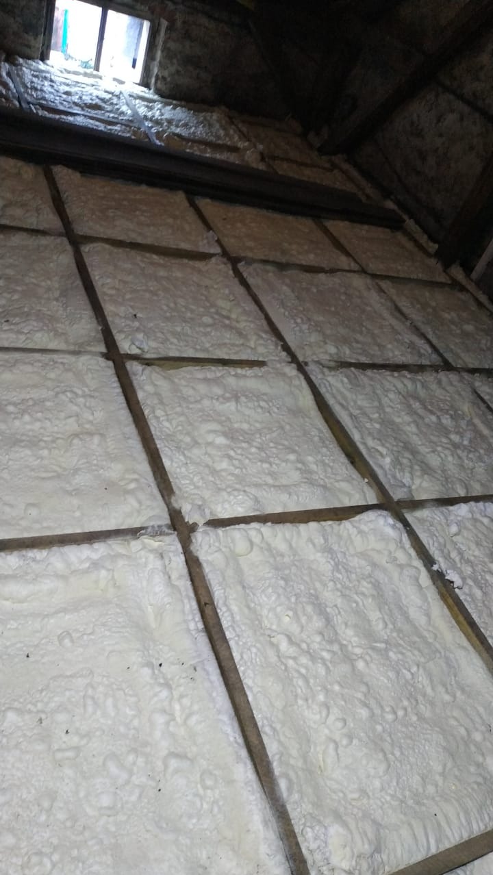 Izolacja poddasza pianą PUR pomiędzy krokwiami, widoczne drewniane elementy konstrukcji dachu i okno dachowe w tle.