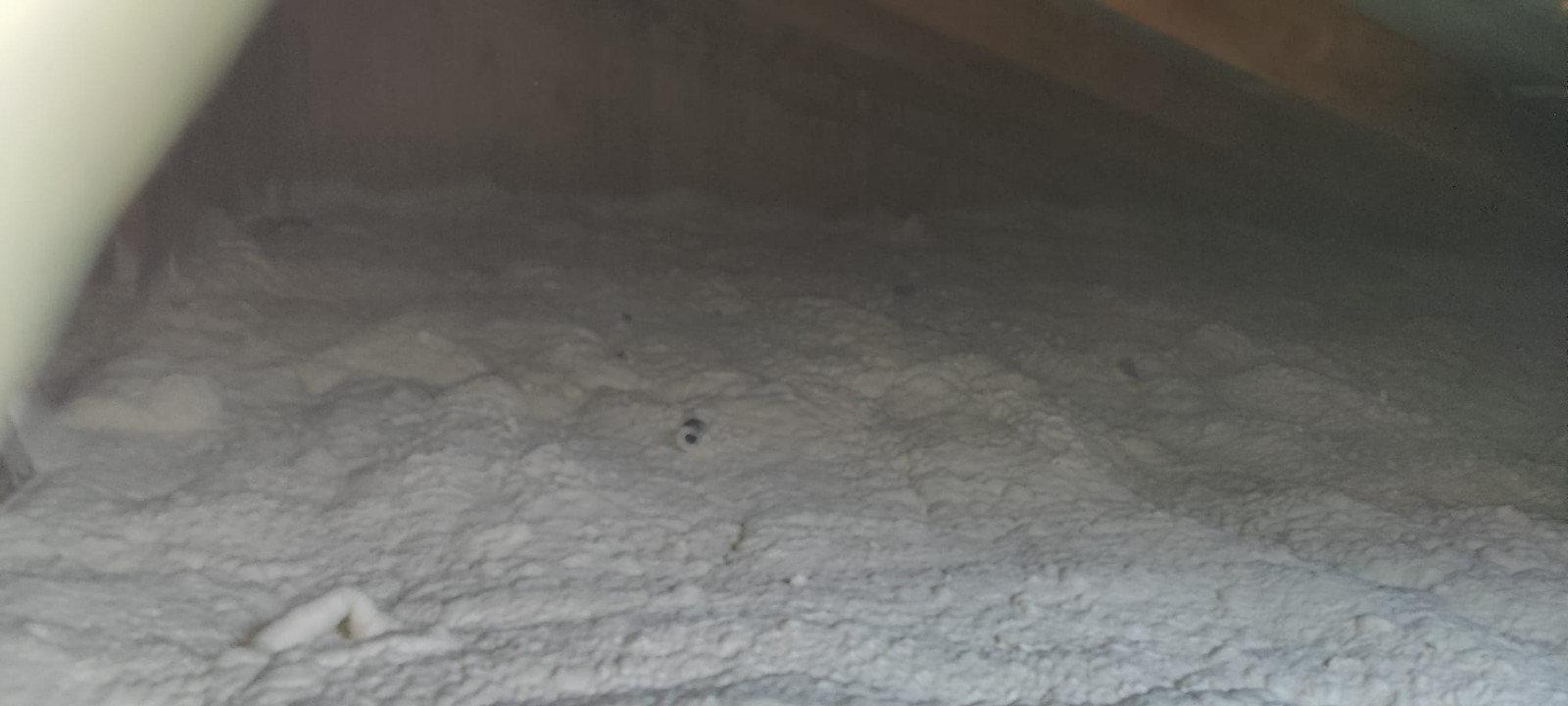 Izolacja poddasza pianką poliuretanową, nierównomierna struktura, widoczne fragmenty konstrukcji dachu, szaro-biała kolorystyka, oświetlenie od góry.