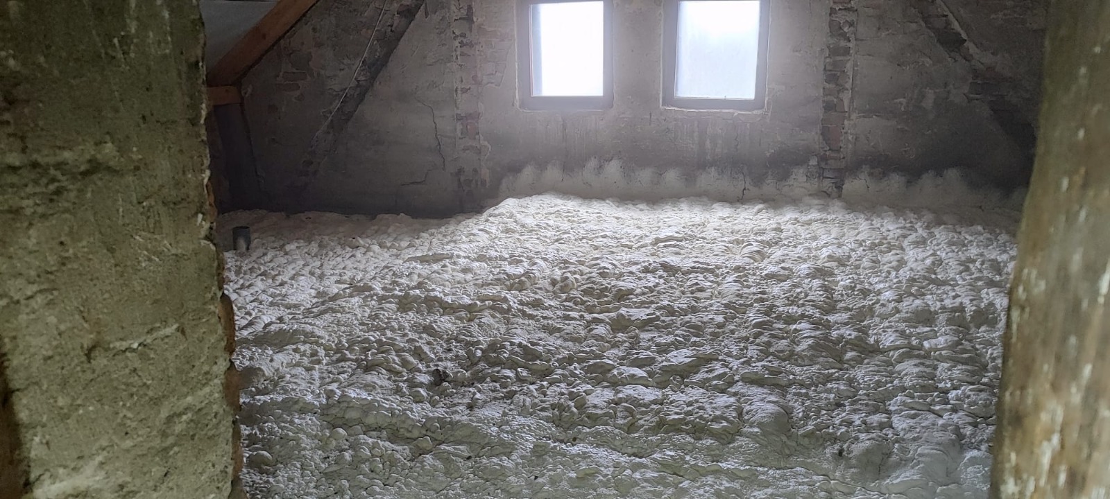 Poddasze po izolacji pianką PUR, widoczne dwie małe okna i fragmenty cegieł. Pianka wypełnia całą przestrzeń, tworząc jednolitą warstwę izolacyjną.