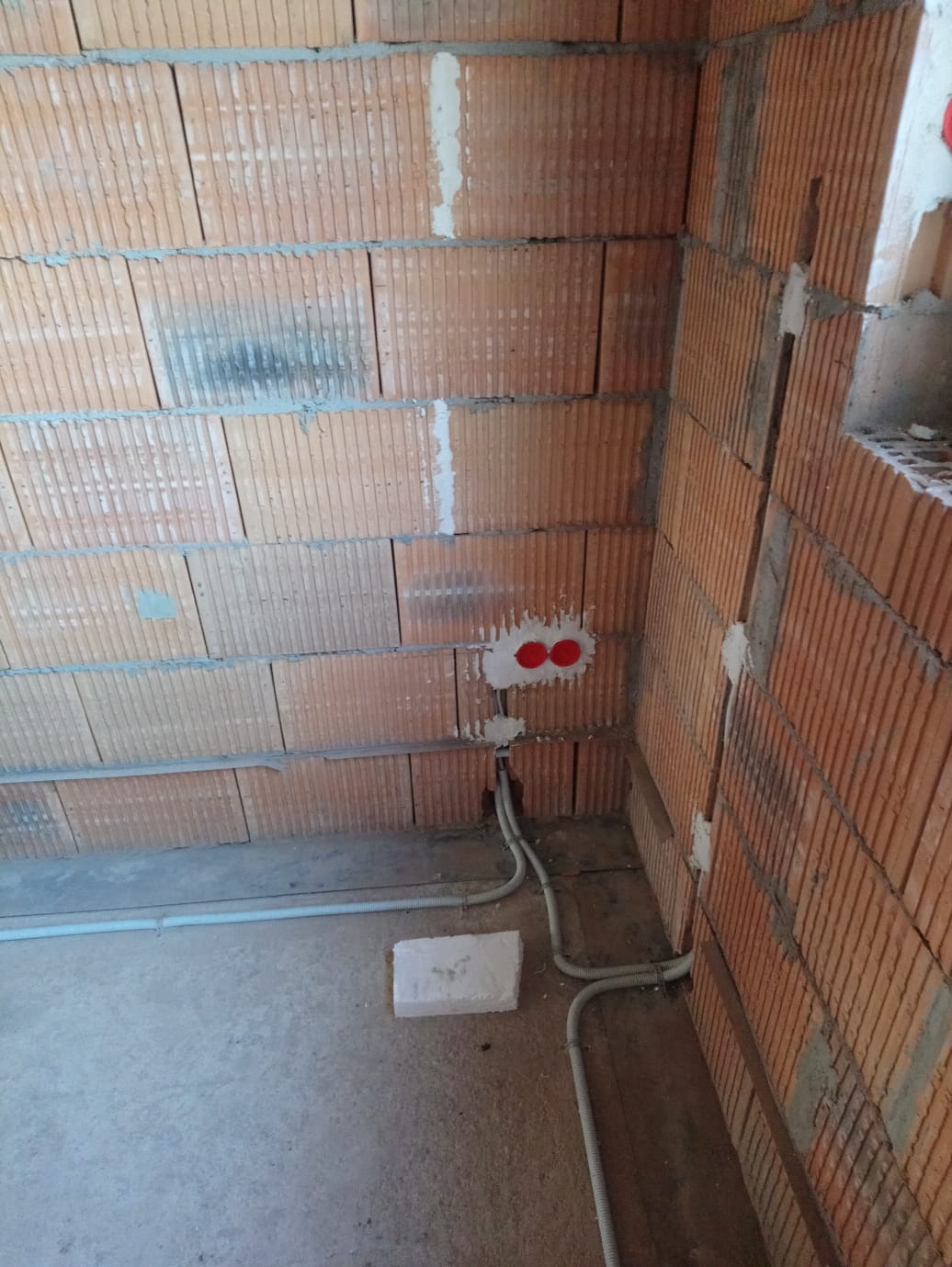 Surowy mur z cegły dziurawki z puszkami elektrycznymi i przewodami w peszlach, widoczne na podłodze. Etap instalacji przed tynkowaniem.