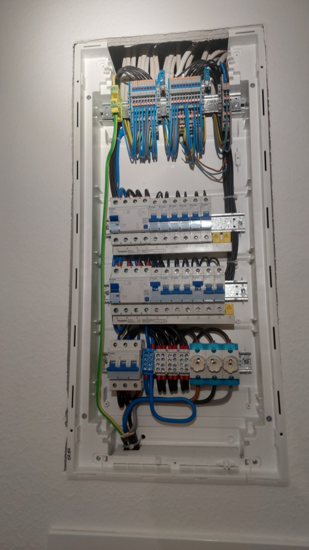 Rozdzielnica elektryczna w białej obudowie, z widocznymi bezpiecznikami, przewodami i złączkami, zamontowana na jasnej ścianie.