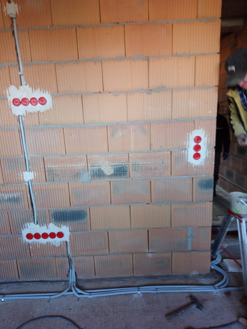 Ściana z cegieł z zamontowanymi puszkami elektrycznymi i przewodami w szarych rurkach. Widoczne czerwone wkładki w puszkach i fragment drabiny.