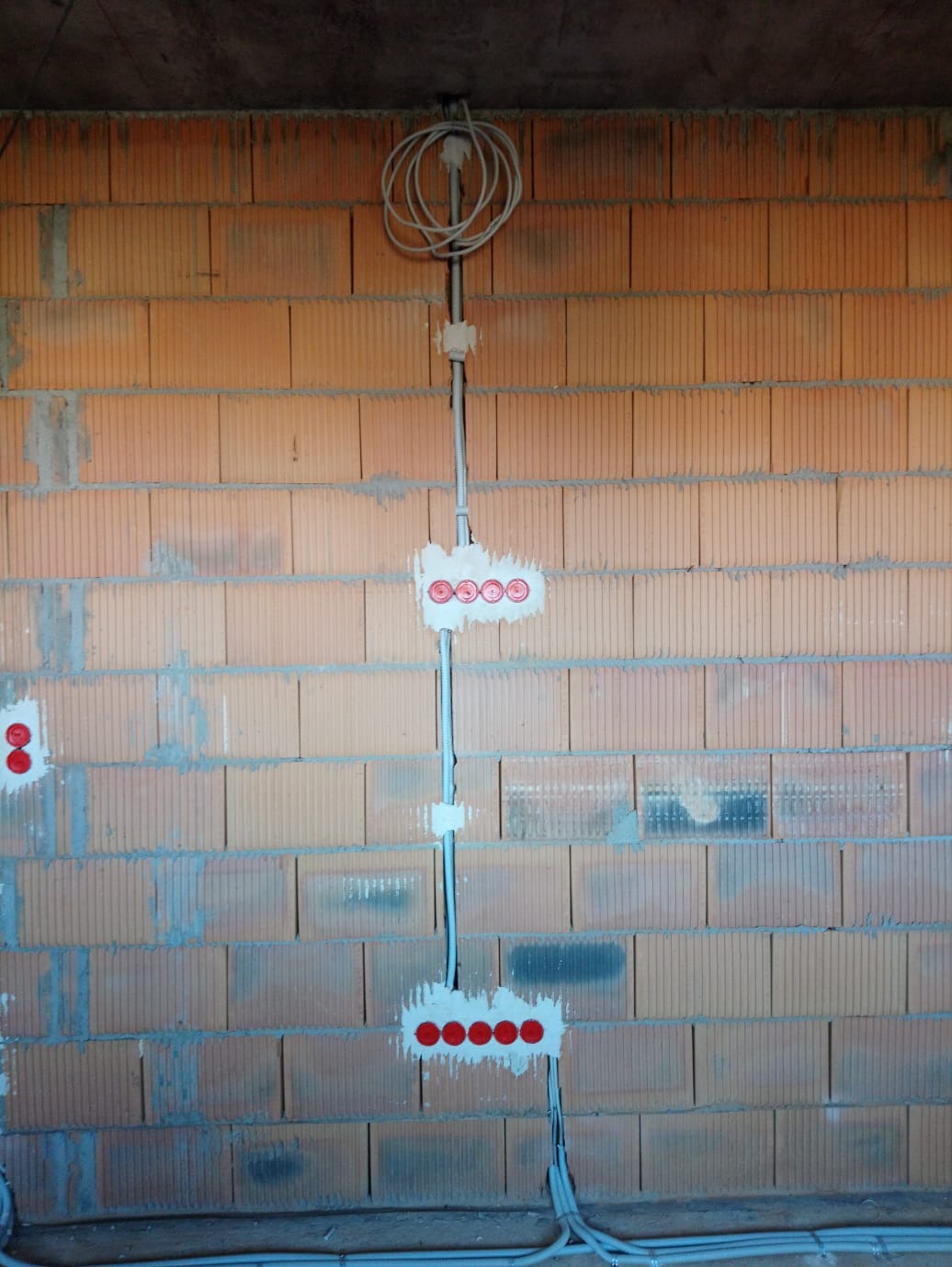 Instalacja elektryczna w ścianie z cegły: przewody, puszki z czerwonymi zaślepkami. Widoczne kable u dołu i zwój kabla pod sufitem. Etap prac instalacyjnych.