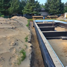 EMEREN SPÓŁKA Z OGRANICZONĄ ODPOWIEDZIALNOŚCIĄ - Fundamenty domu w trakcie budowy: izolacja pionowa z folii kubełkowej, zbrojenie wystające z betonu, wykop z piaskiem. W tle drzewa i częściowo widoczny budynek.