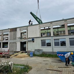 EMEREN SPÓŁKA Z OGRANICZONĄ ODPOWIEDZIALNOŚCIĄ - Budowa budynku z pustaków: robotnicy na dachu montują zieloną konstrukcję stalową za pomocą dźwigu. Widoczny kontener na gruz i elementy konstrukcyjne.