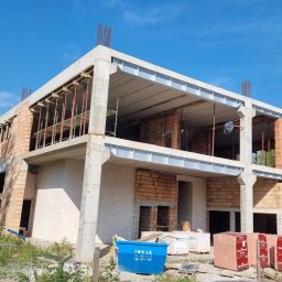 EMEREN SPÓŁKA Z OGRANICZONĄ ODPOWIEDZIALNOŚCIĄ - Dwukondygnacyjny budynek w trakcie budowy, z widocznymi elementami konstrukcyjnymi i częściowo wymurowanymi ścianami z cegły, na tle zieleni i błękitnego nieba.