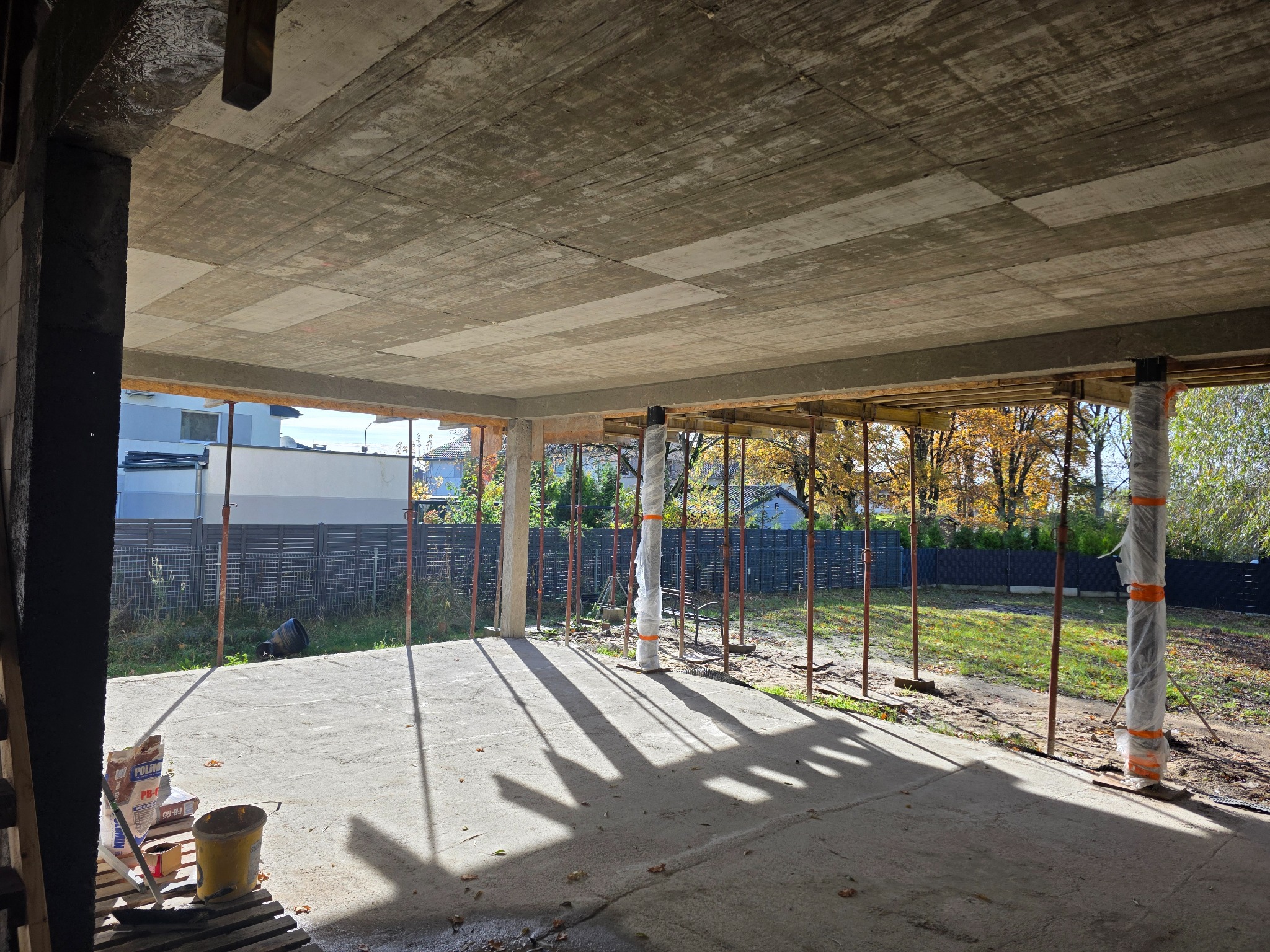 Surowy strop żelbetowy z deskowaniem i podporami, widok z wnętrza budynku w trakcie budowy. Na podłodze cień podpór, w tle widoczny ogród i zabudowa jednorodzinna.