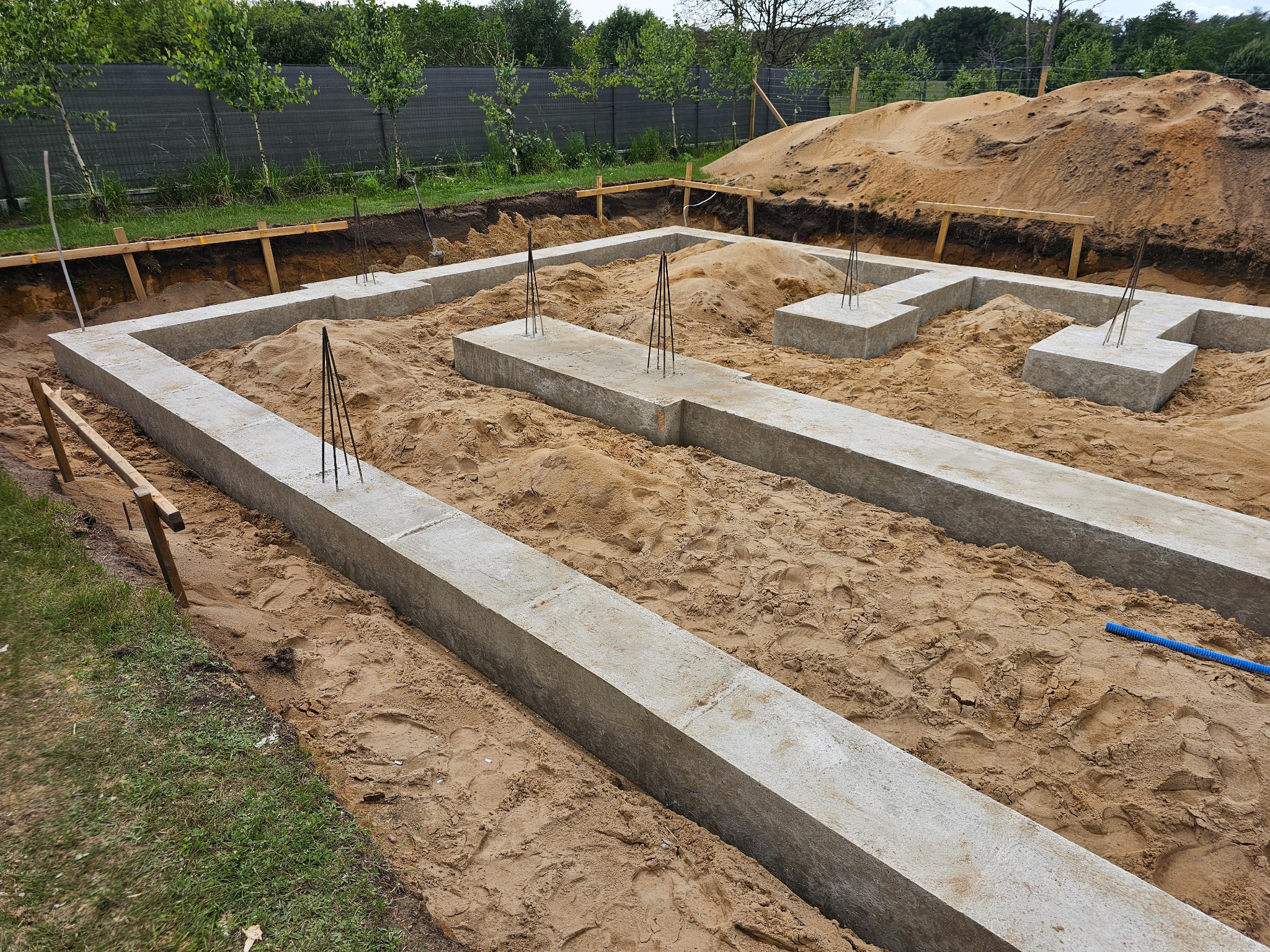 Zalane betonem fundamenty domu jednorodzinnego z wystającymi prętami zbrojeniowymi, otoczone piaskiem i deskowaniem. Widok z góry na wykop pod budowę.