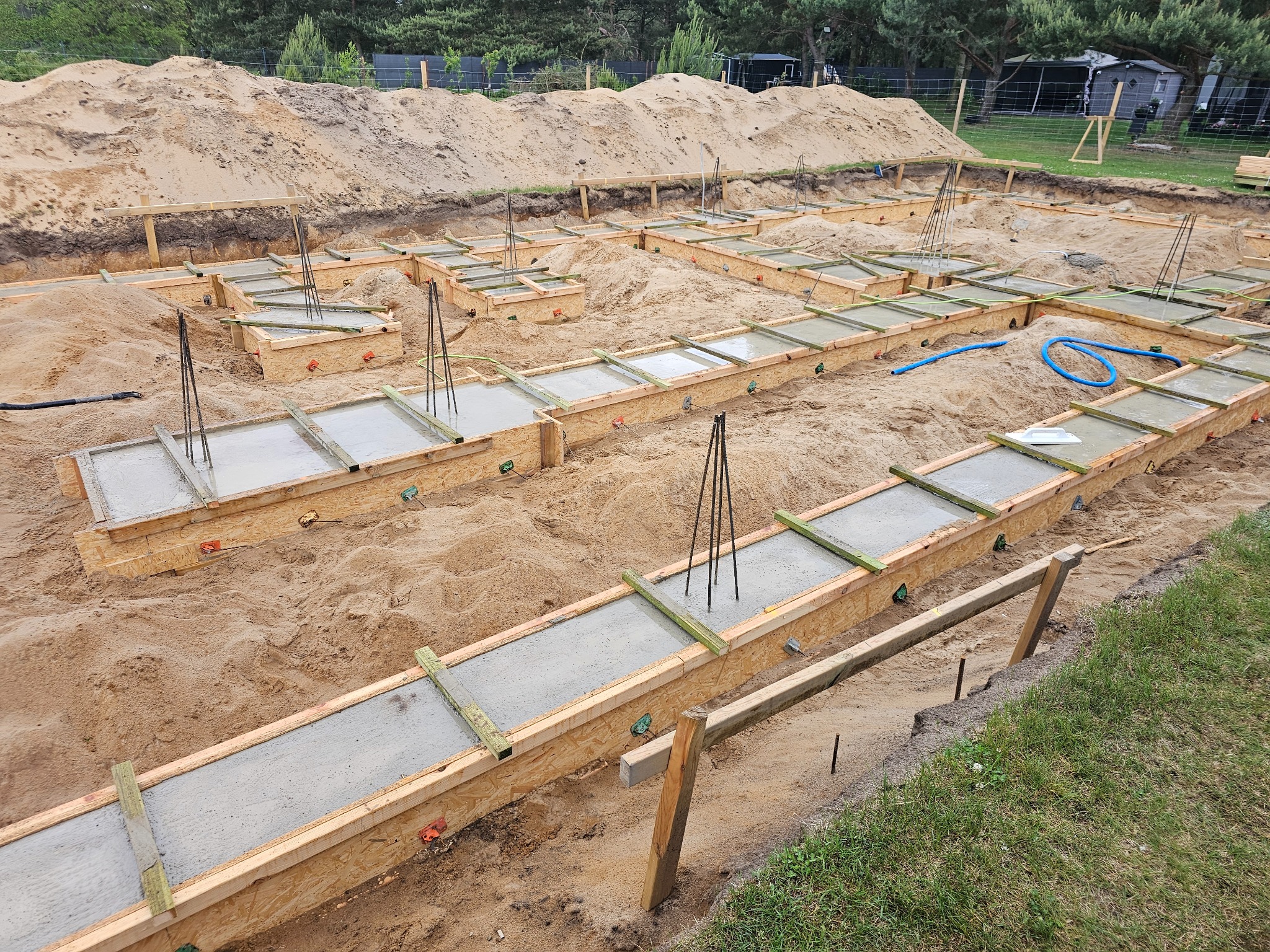 Zalane betonem szalunki fundamentowe na piaszczystym podłożu, widoczne wystające pręty zbrojeniowe i niebieski wąż. W tle usypana ziemia i drzewa.