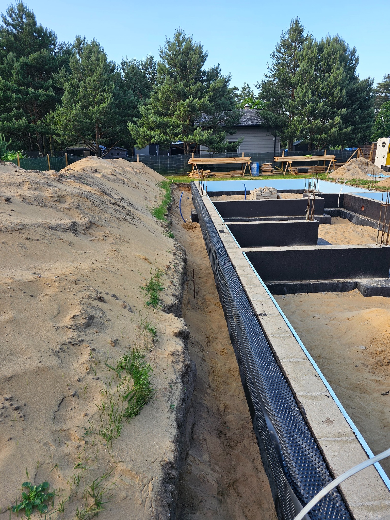 Fundamenty domu w trakcie budowy: izolacja pionowa z folii kubełkowej, zbrojenie wystające z betonu, wykop z piaskiem. W tle drzewa i częściowo widoczny budynek.