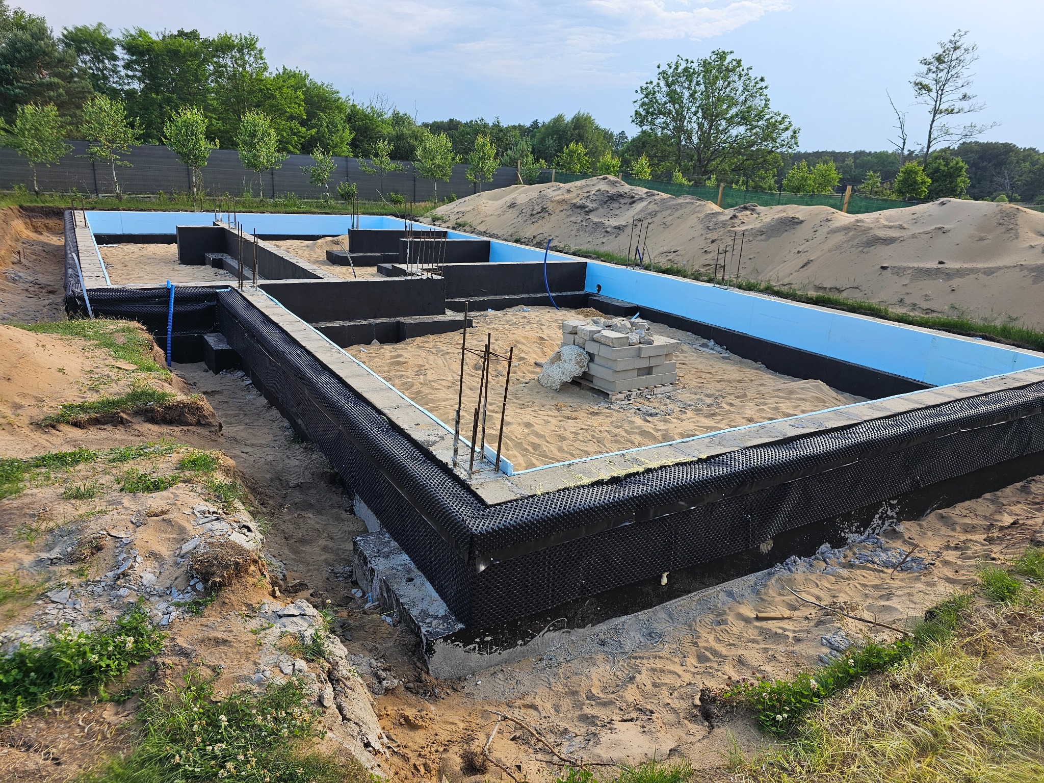 Fundamenty domu w trakcie budowy: izolacja termiczna, zbrojenie, piasek. Widok z góry na wykop z przygotowanymi ławami fundamentowymi i izolacją.