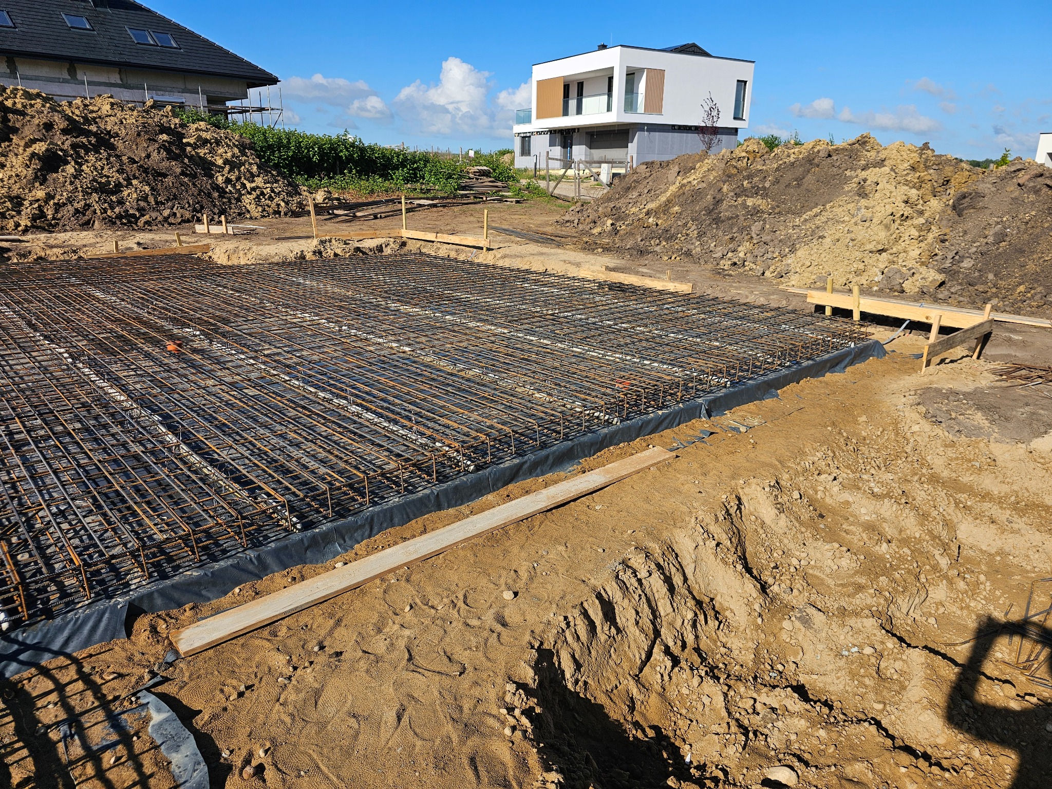 Zbrojenie fundamentu pod budowę domu jednorodzinnego. Widoczne ułożone pręty zbrojeniowe na czarnej folii, drewniane szalunki i usypana ziemia w tle.