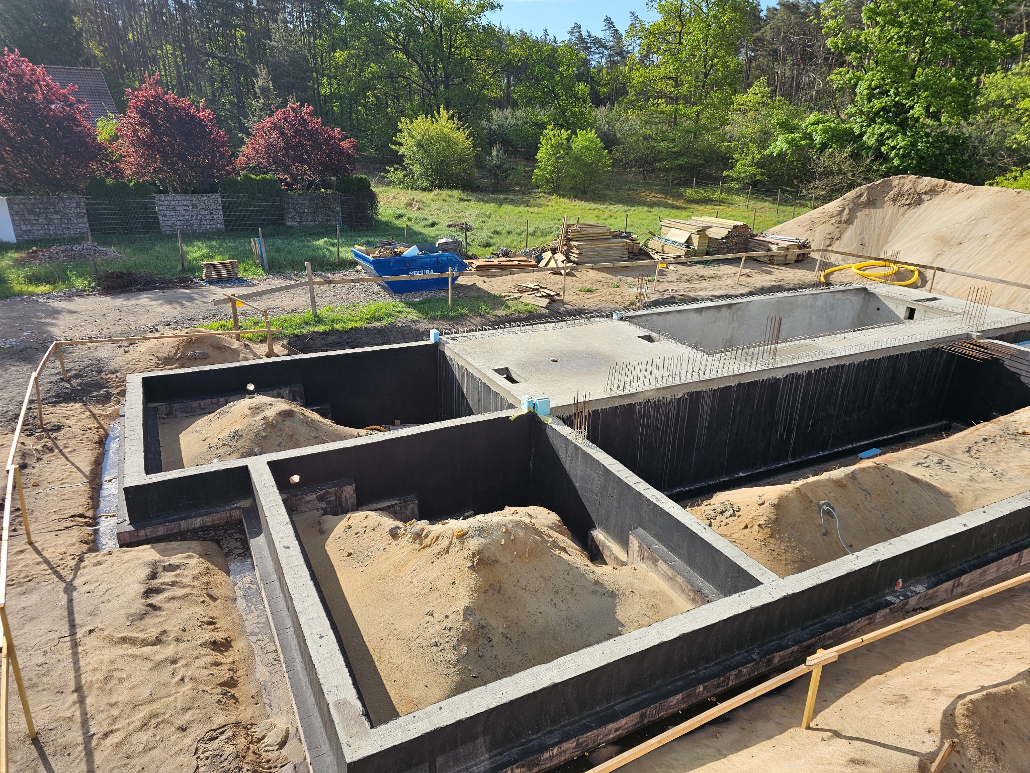 Zalane fundamenty domu z hydroizolacją i zbrojeniami, widok z góry na wykop z piaskiem, w tle drzewa i niebieski kontener budowlany.