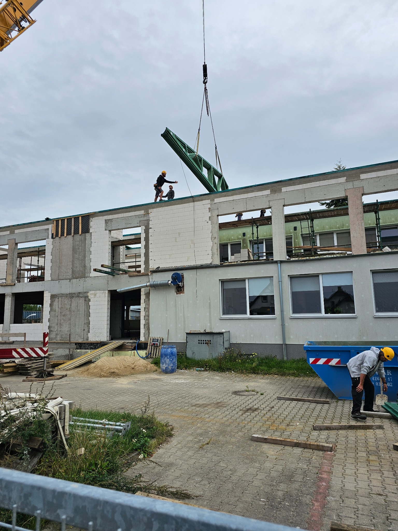 Budowa budynku z pustaków: robotnicy na dachu montują zieloną konstrukcję stalową za pomocą dźwigu. Widoczny kontener na gruz i elementy konstrukcyjne.