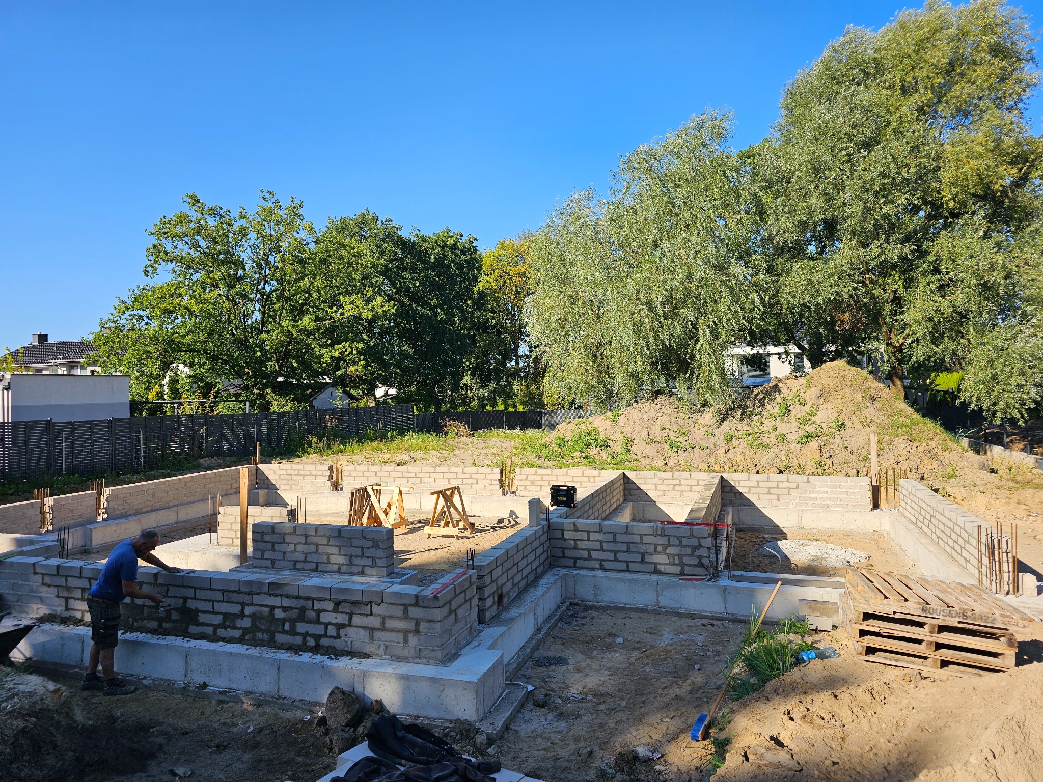 Fundamenty domu w trakcie budowy: murarz w niebieskiej koszulce układa bloczki, widoczne zbrojenia, paleta i drzewa w tle pod błękitnym niebem.