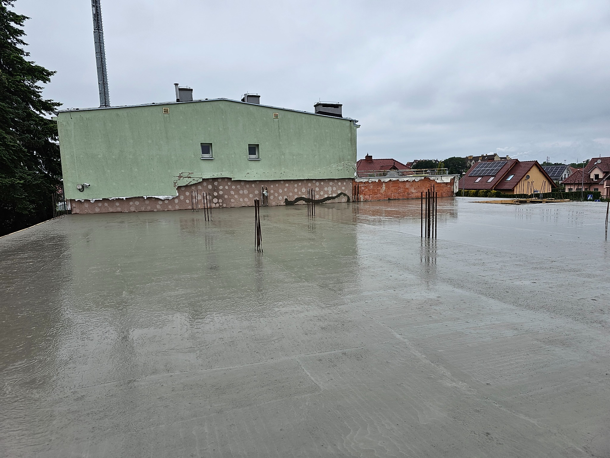 Świeżo wylany, mokry strop betonowy z wystającymi prętami zbrojeniowymi na budowie domu jednorodzinnego w pochmurny dzień. Widoczne sąsiednie budynki.
