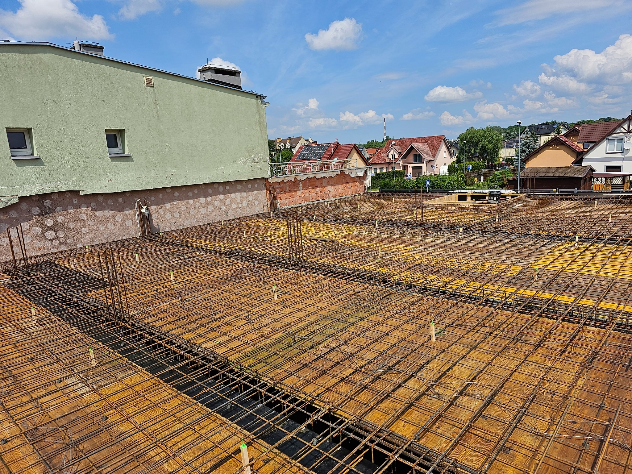 Zbrojenie stropu budynku wielorodzinnego z widocznymi deskami szalunkowymi i wystającymi prętami zbrojeniowymi. W tle zabudowa jednorodzinna i błękitne niebo.