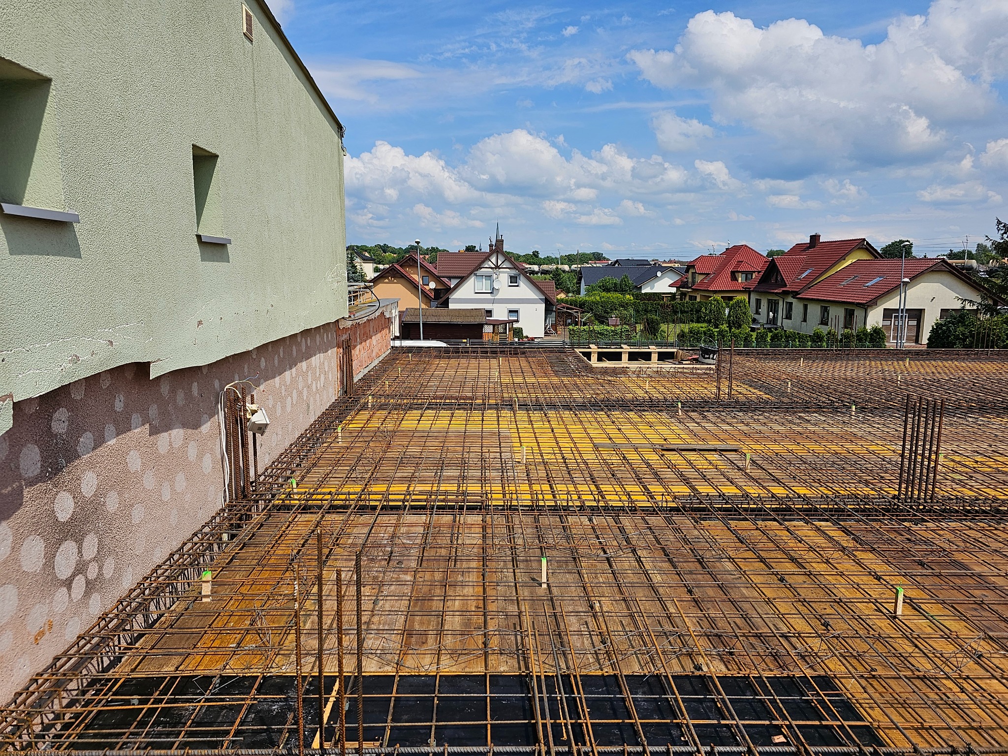 Zbrojenie stropu budynku w trakcie budowy, widok z góry na siatkę z prętów stalowych, w tle zabudowa jednorodzinna i błękitne niebo z chmurami.
