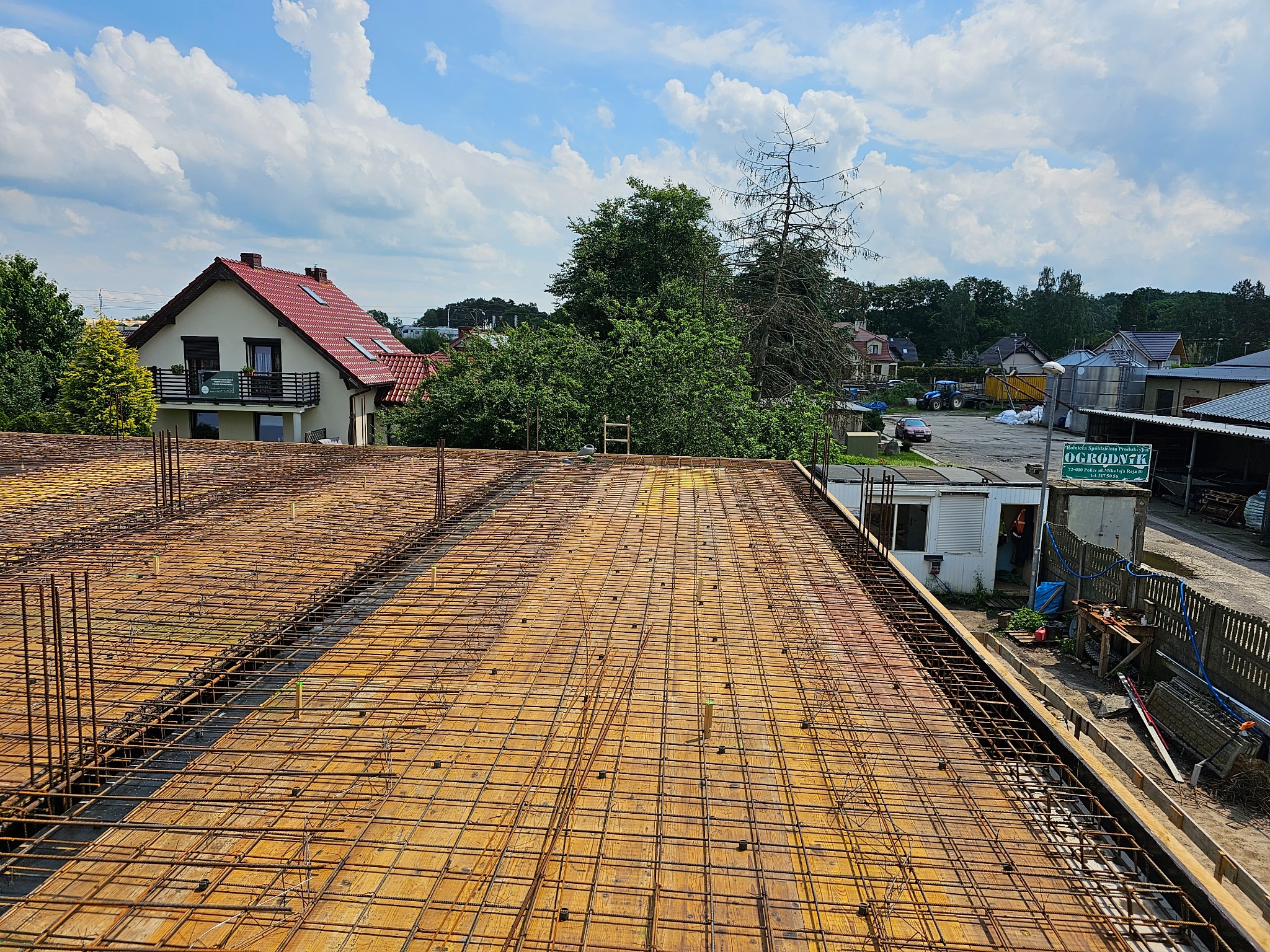 Zbrojenie stropu budynku jednorodzinnego z widoczną siatką zbrojeniową i deskowaniem, w tle zabudowa wiejska i błękitne niebo z chmurami.