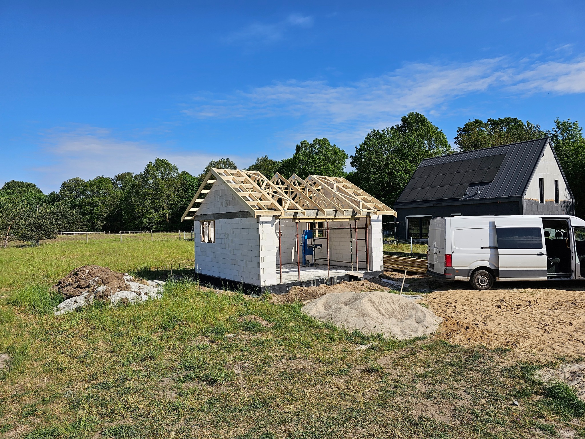 Budowa domu z białych bloczków: ściany wzniesione, drewniana konstrukcja dachu w trakcie montażu, na tle zieleni i nowoczesnego domu z panelami słonecznymi.