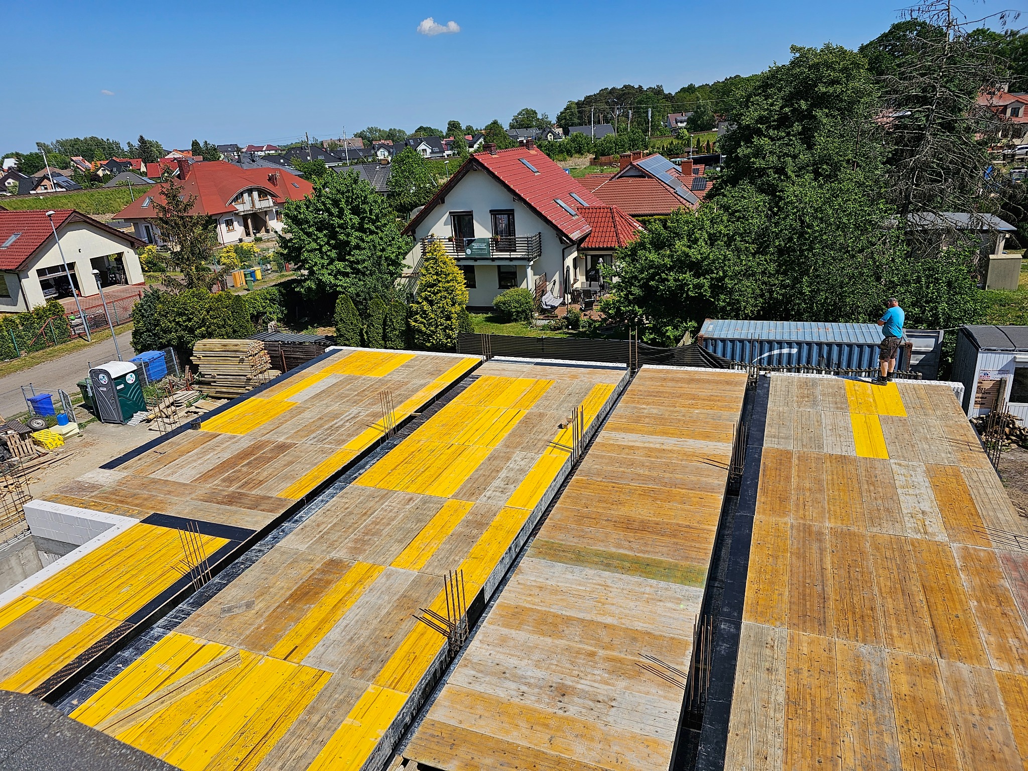Z góry: Szalunek stropu z żółtymi elementami na budowie domu jednorodzinnego w słoneczny dzień. W tle zabudowa mieszkalna i zieleń.